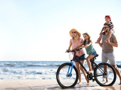 A happy family of four enjoying a sunny day on the Ocean City, Maryland shore, with a mother and daughter on a bicycle and a father carrying his son on his shoulders along the water's edge.